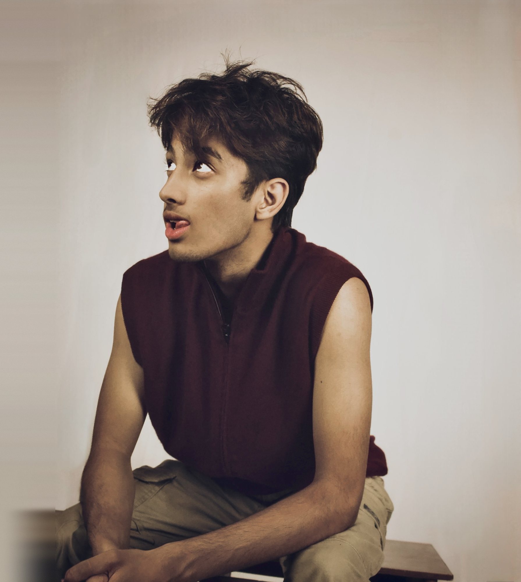 A young man in a maroon knitted vest looking up, seated against a pale studio wall